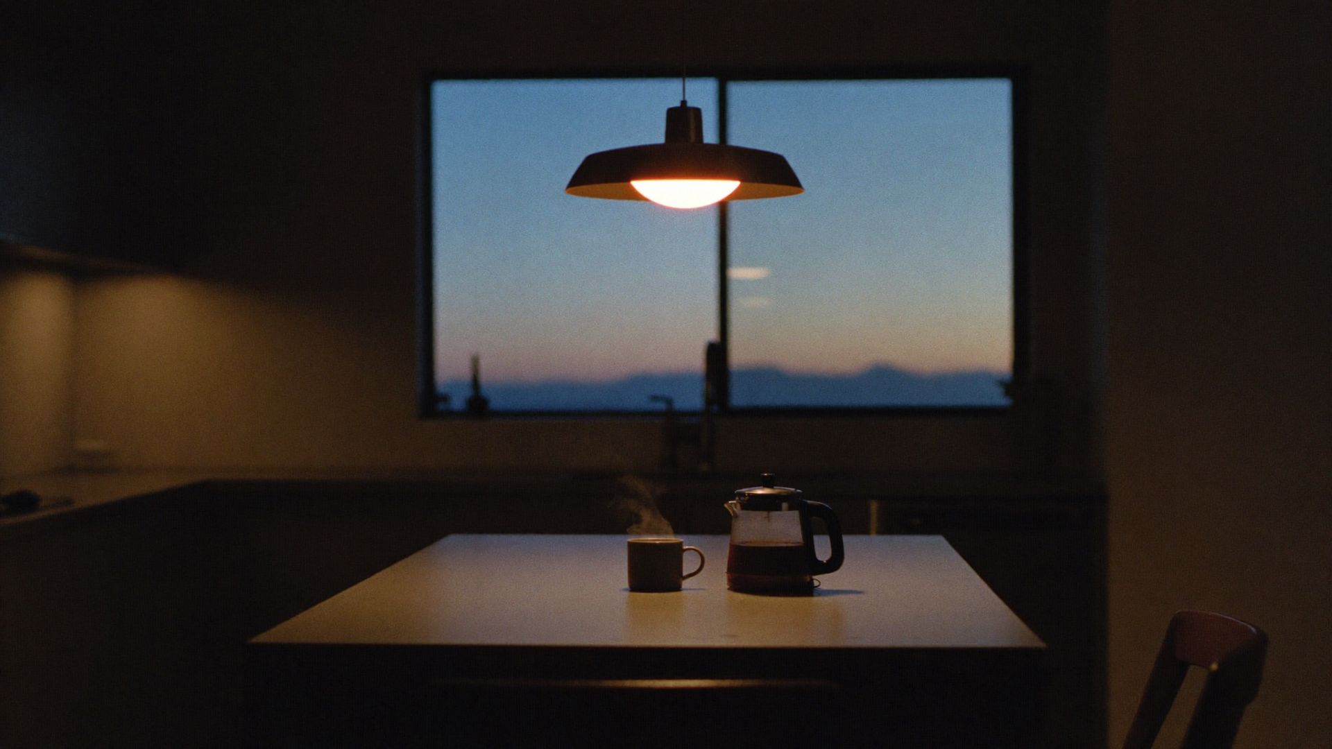 A dark kitchen before sunrise, one pendant lamp over the island, coffee kettle and mug, hills in the window.