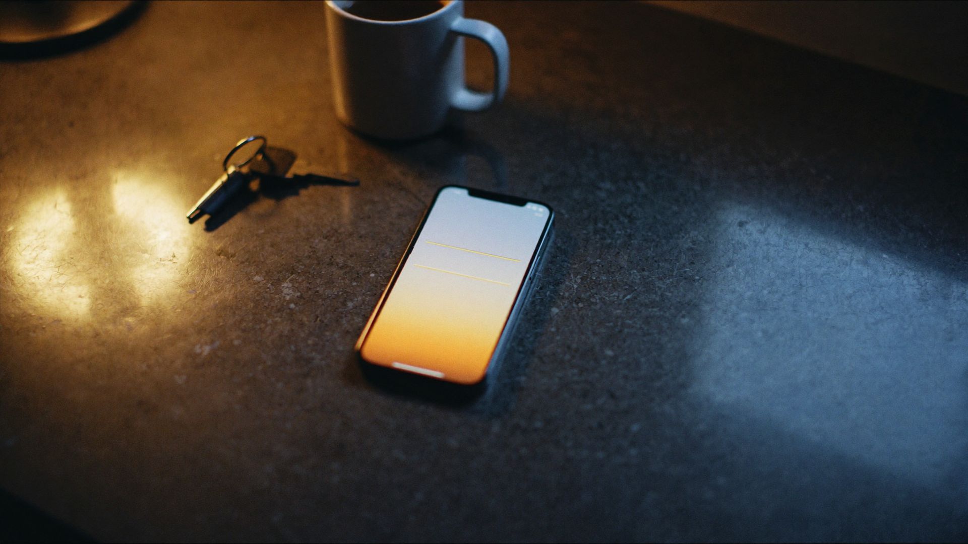 A phone face up on a kitchen counter next to a coffee mug and house keys.